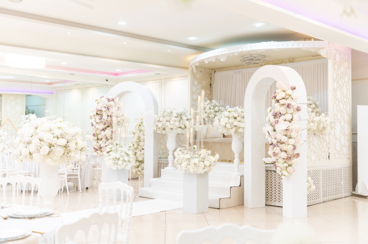 Salle de réception décorée avec des arches blanches ornées de fleurs et un podium entouré de compositions florales.