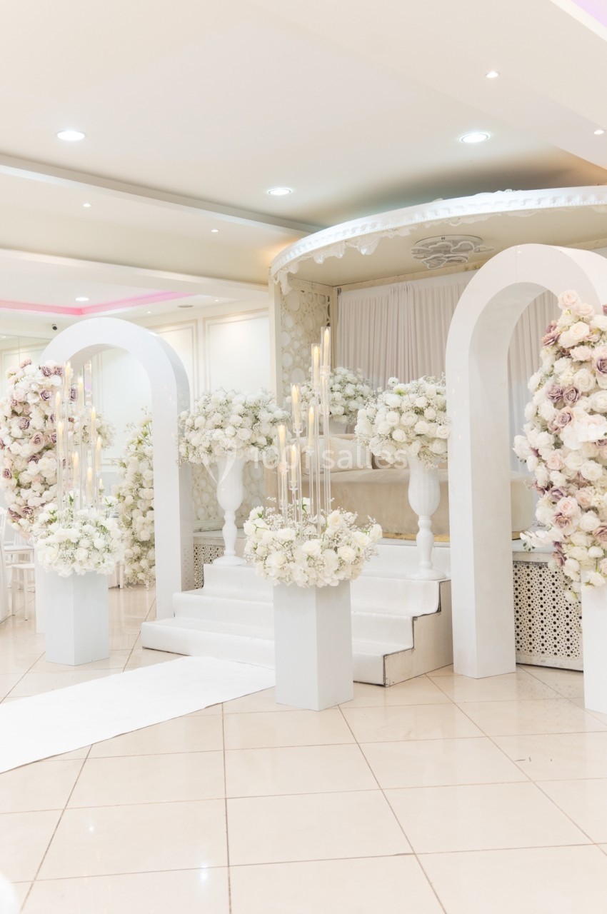 Décoration de mariage avec arche blanche, fleurs blanches et roses, chandeliers et estrade dans une salle lumineuse.