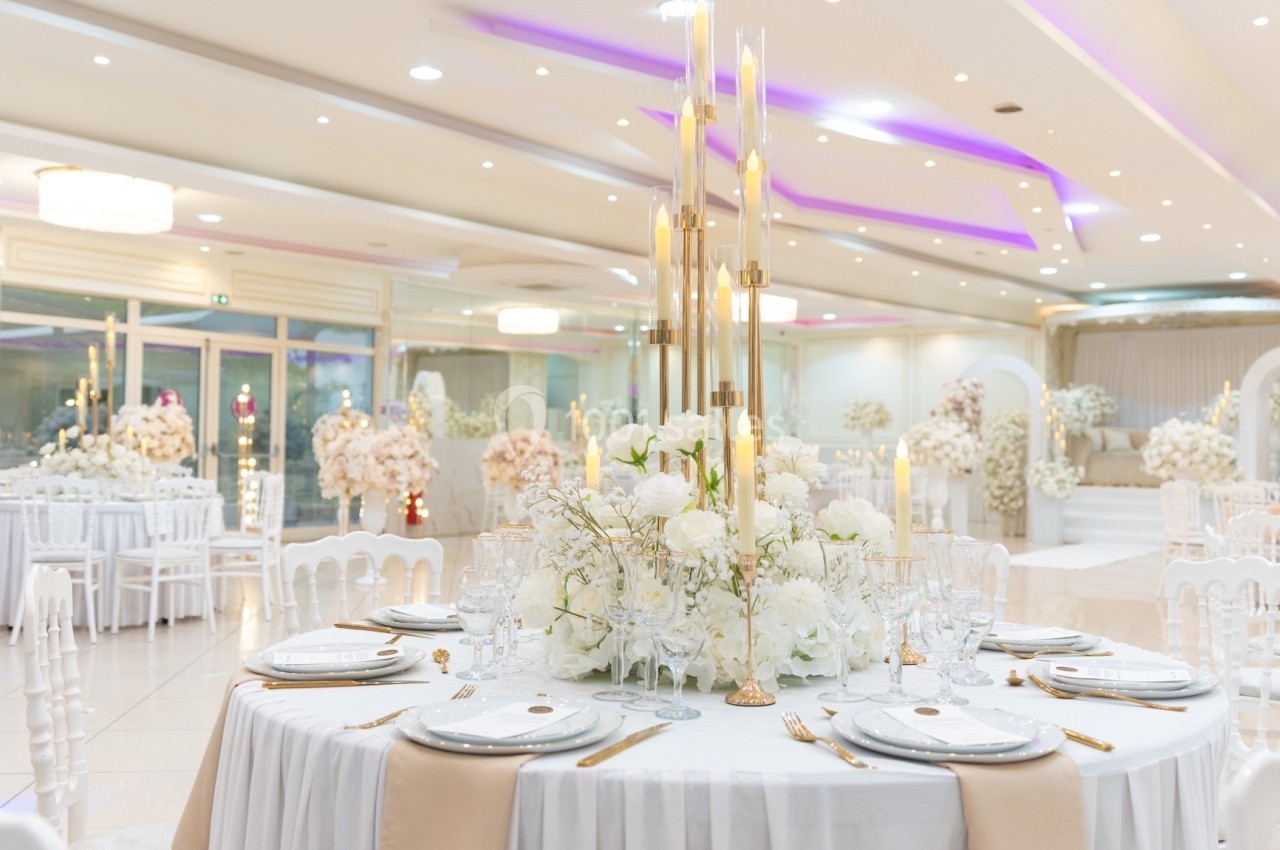 Salle de réception élégante avec tables décorées de fleurs blanches, vaisselle raffinée et chandeliers dorés.