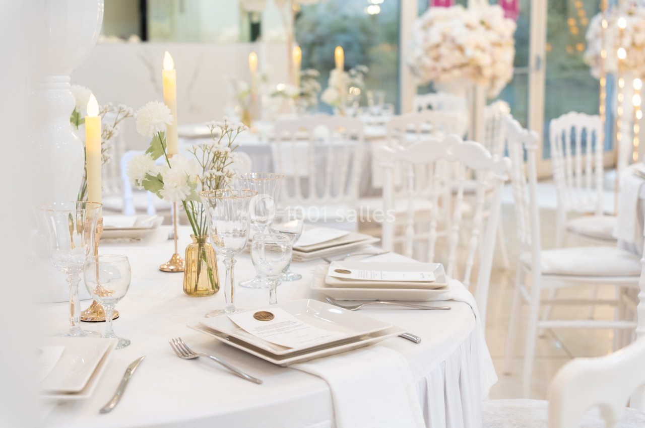 Salle de réception élégante avec tables décorées de fleurs blanches, bougies et vaisselle raffinée.