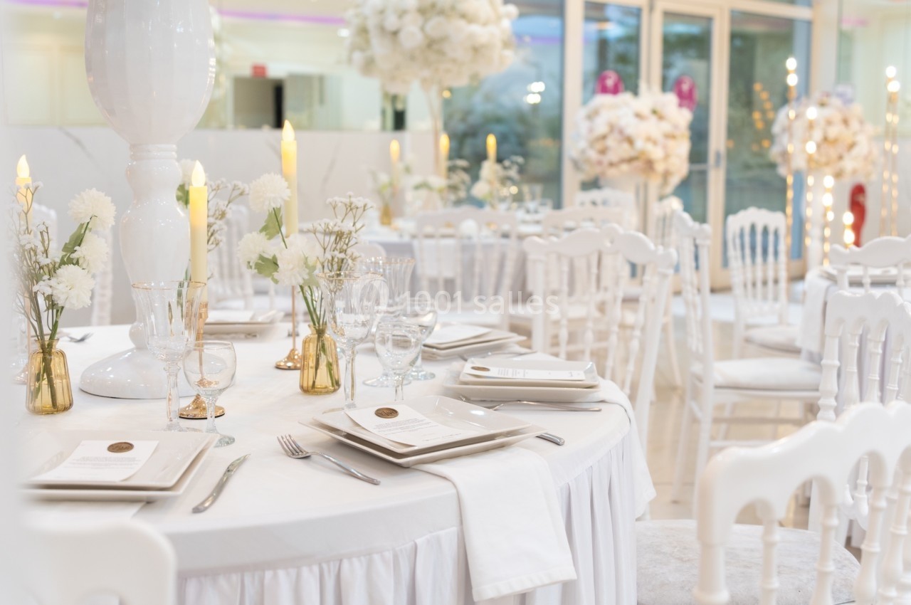 Salle de réception élégante avec tables rondes dressées, décorations florales blanches et bougies allumées.