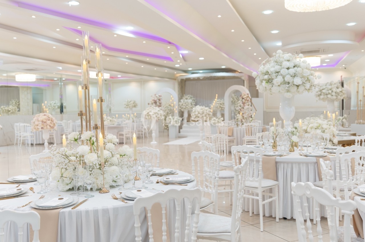 Salle de réception élégante avec tables décorées de fleurs blanches et chaises blanches, éclairage doux et arches fleuries.