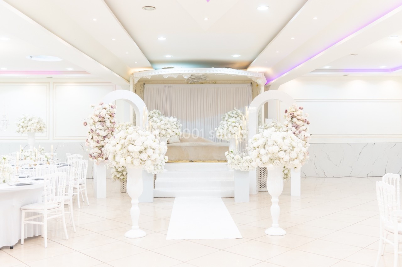 Salle de réception lumineuse décorée de fleurs blanches, avec une scène centrale et des tables élégamment dressées.