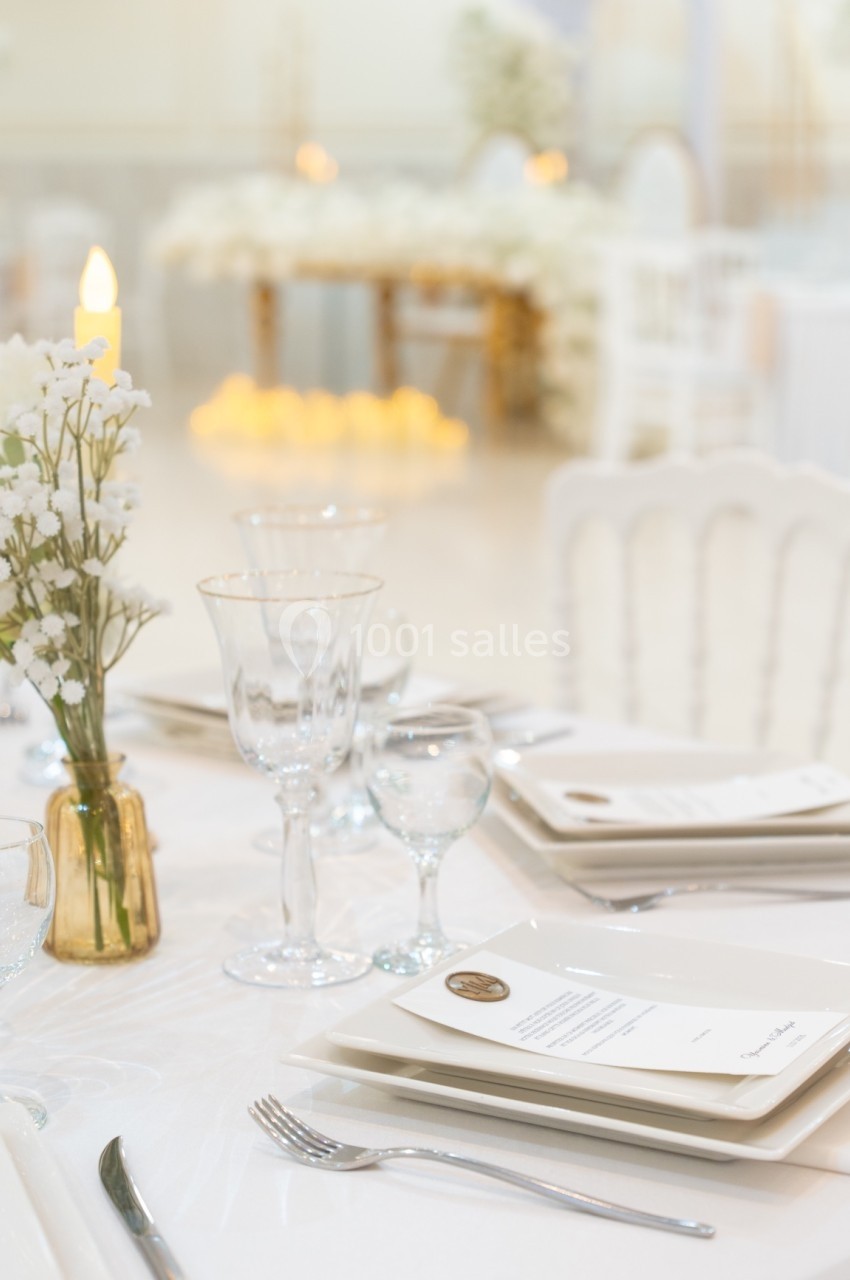 Table élégamment dressée avec vaisselle blanche, verres, menus et vase de fleurs blanches dans une salle lumineuse.