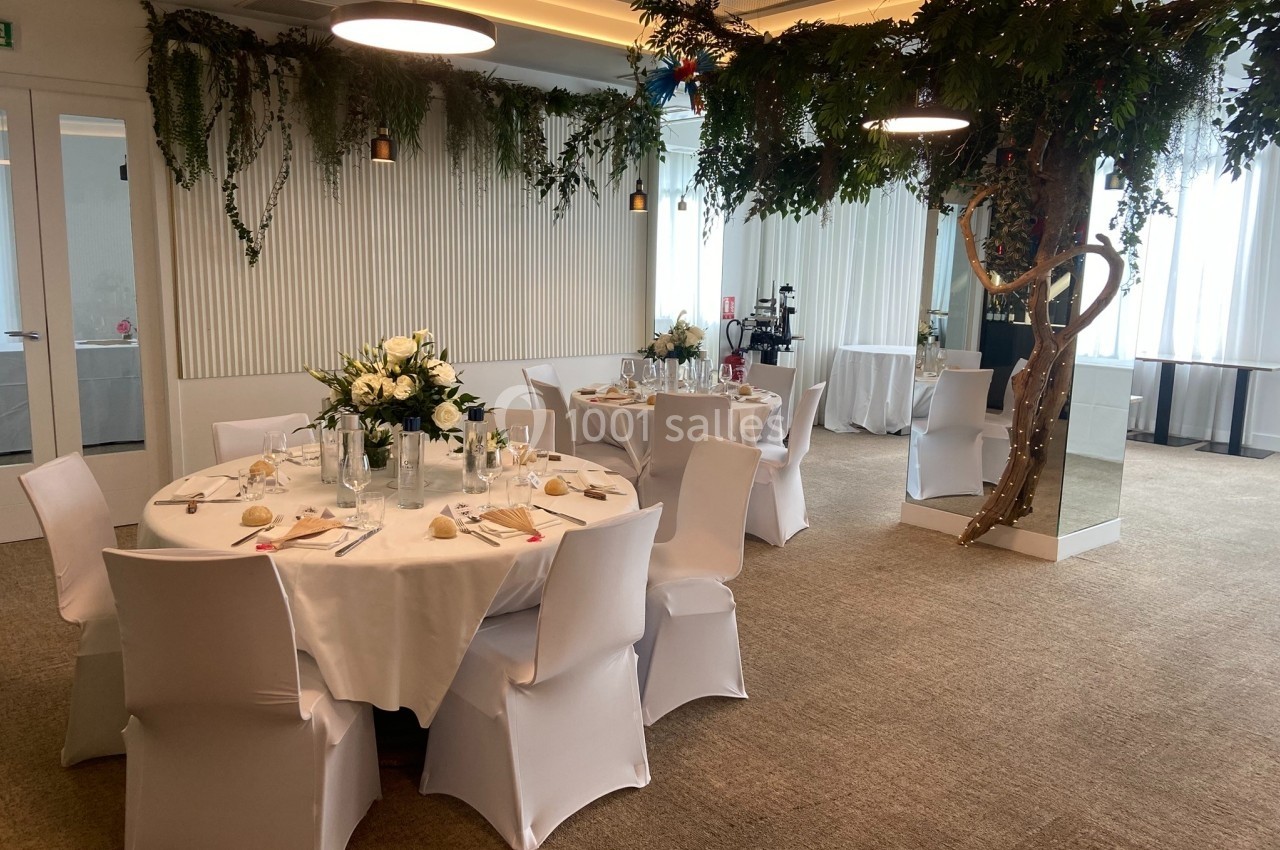 Salle de réception décorée avec des tables rondes dressées, des chaises blanches et des plantes suspendues.