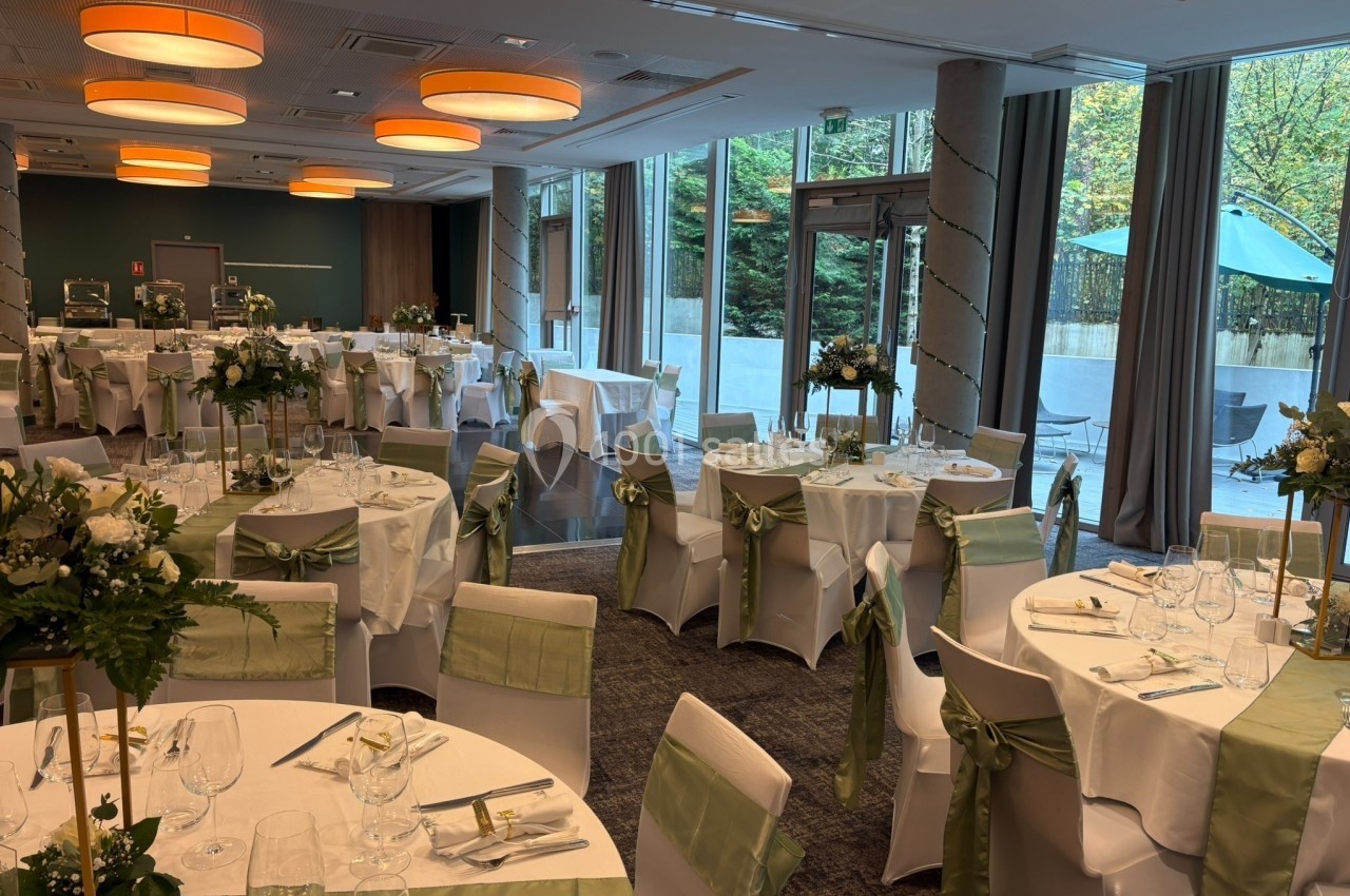 Salle de réception décorée pour un événement, avec tables rondes dressées, nappes blanches et accents verts.