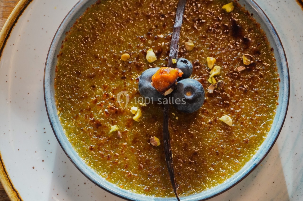 Crème brûlée dorée garnie de myrtilles, d'une noisette, d'éclats de pistache et d'une gousse de vanille.