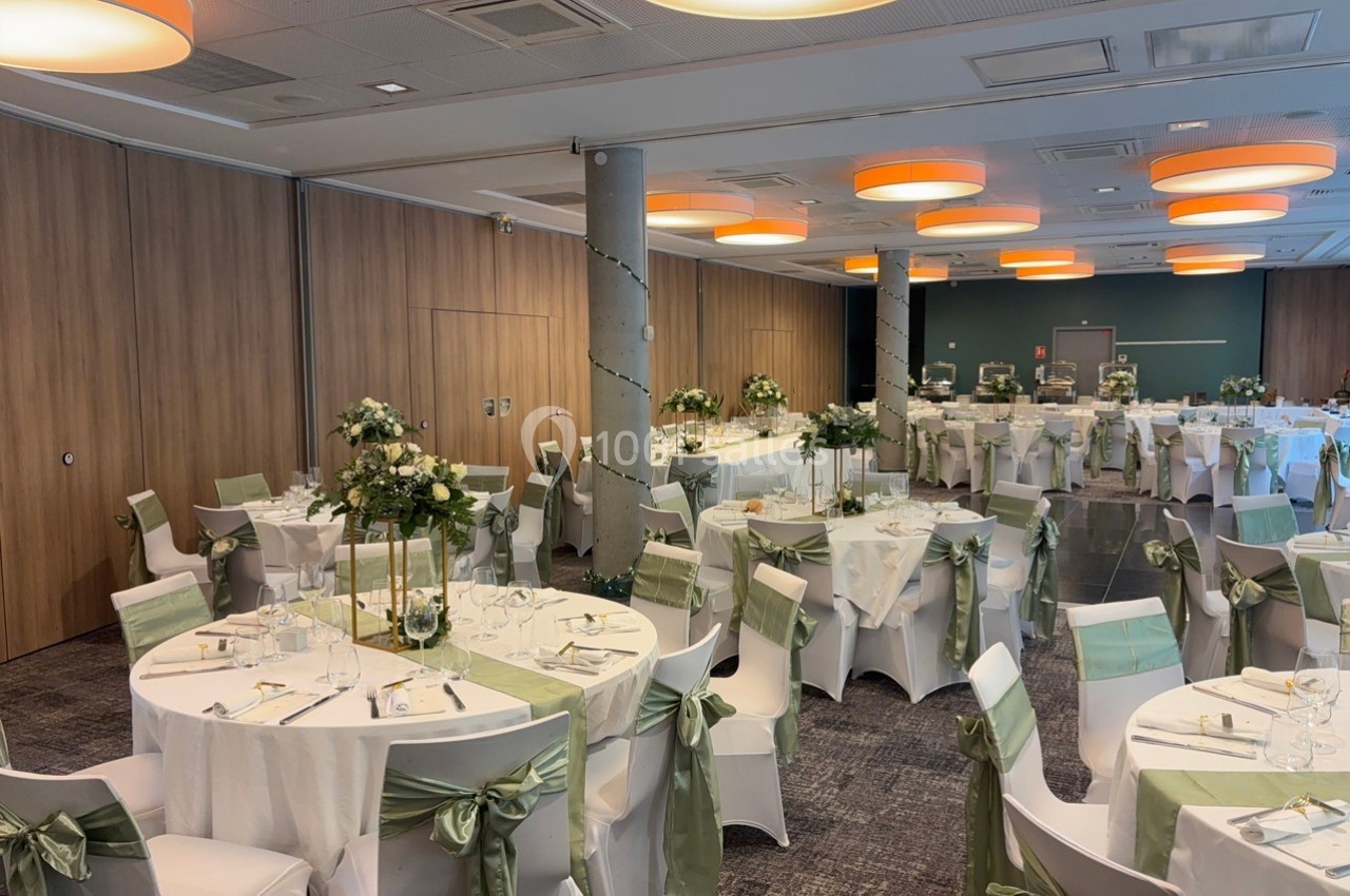 Salle de réception décorée avec des tables rondes, nappes blanches, chaises ornées de rubans verts et centres de table…
