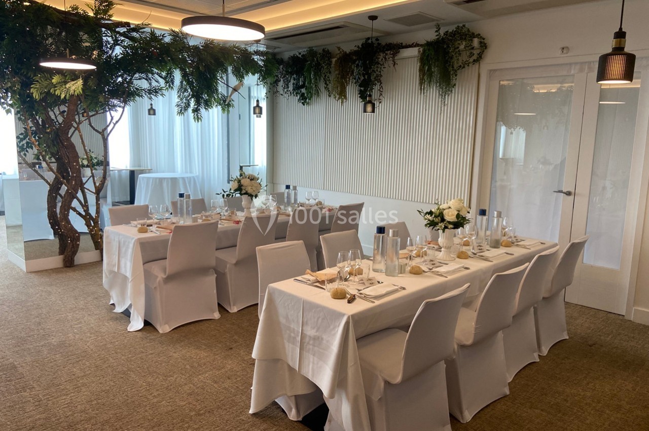 Salle de réception lumineuse avec deux tables dressées, nappes blanches et décorations florales élégantes.