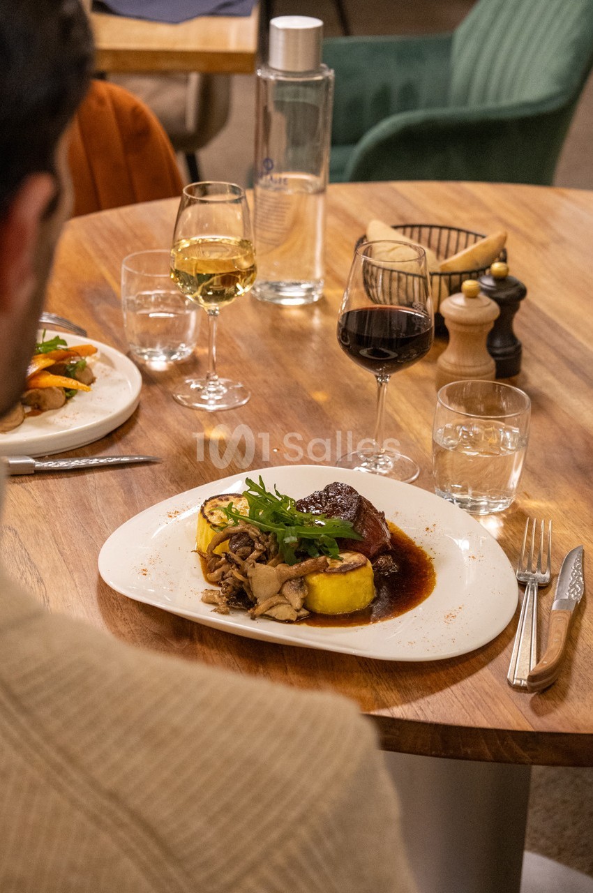 Assiette de viande avec sauce, polenta, champignons et roquette, servie sur une table avec verres de vin et eau.