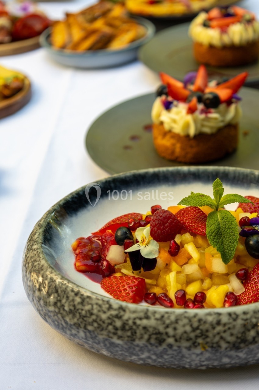 Salade de fruits frais garnie de menthe, avec des desserts et des frites en arrière-plan sur une table dressée.