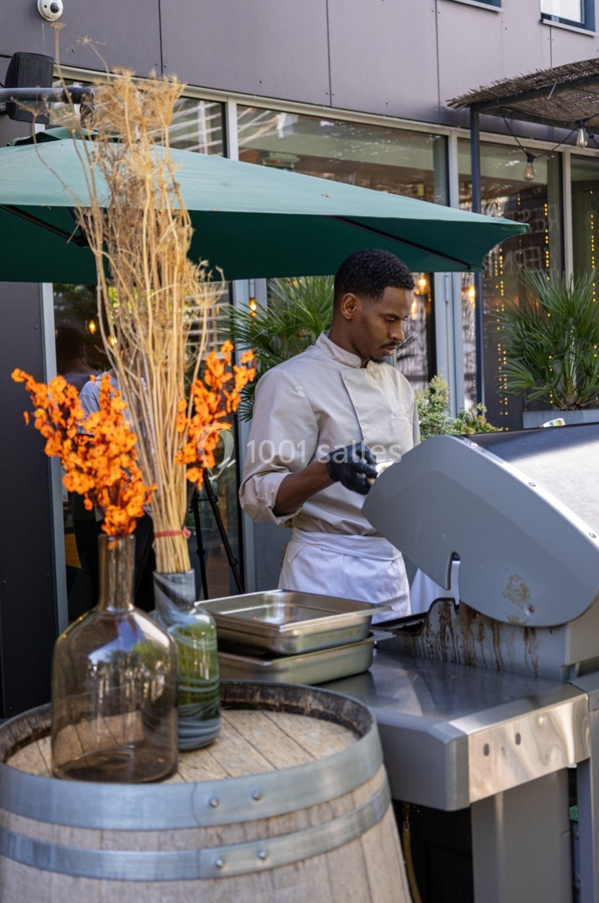 Un chef cuisine sur un barbecue en extérieur, près d'une décoration avec des fleurs séchées et des plantes.