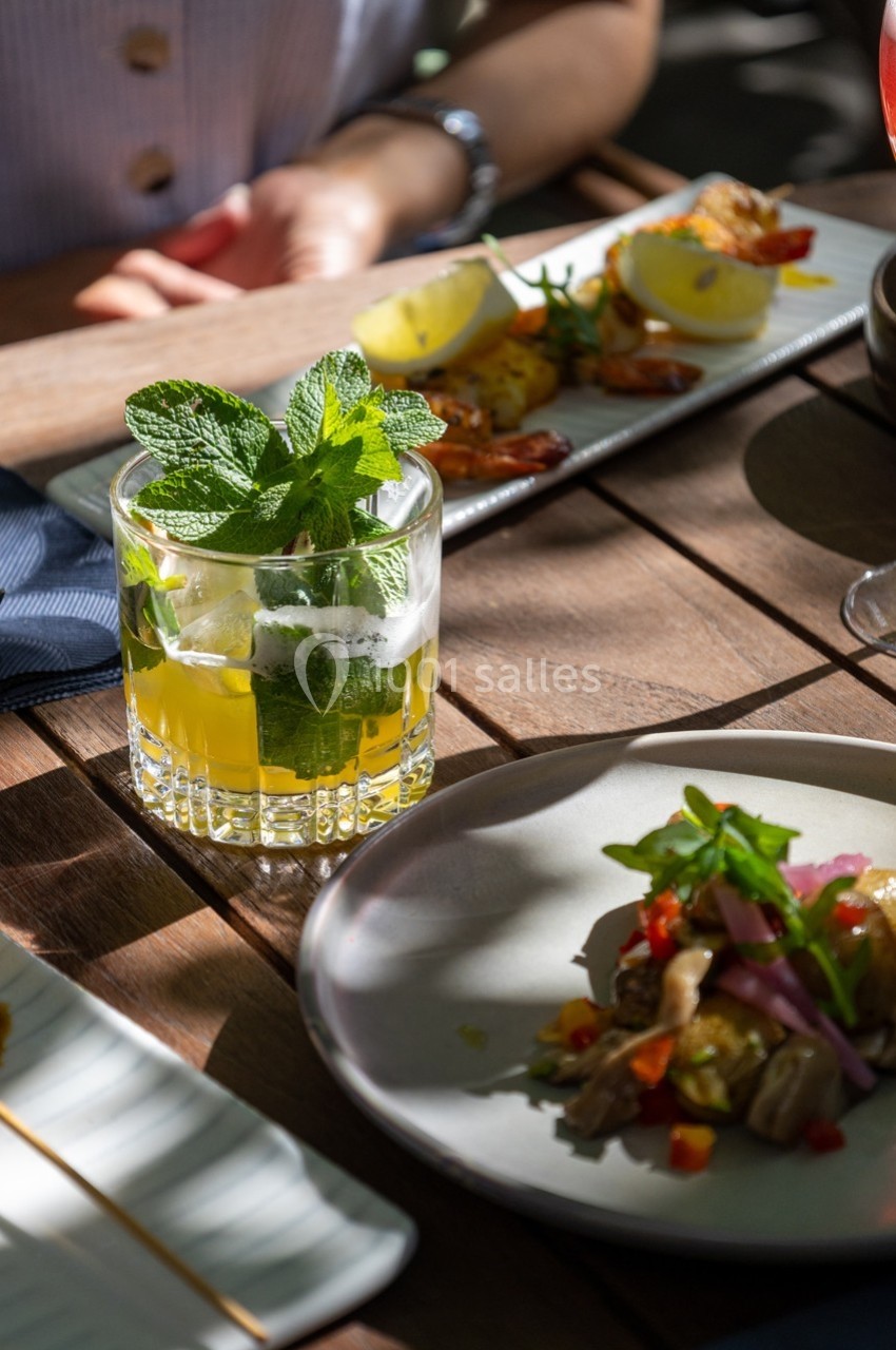 Verre de cocktail à la menthe sur une table en bois, accompagné de plats variés et éclairé par une lumière naturelle.