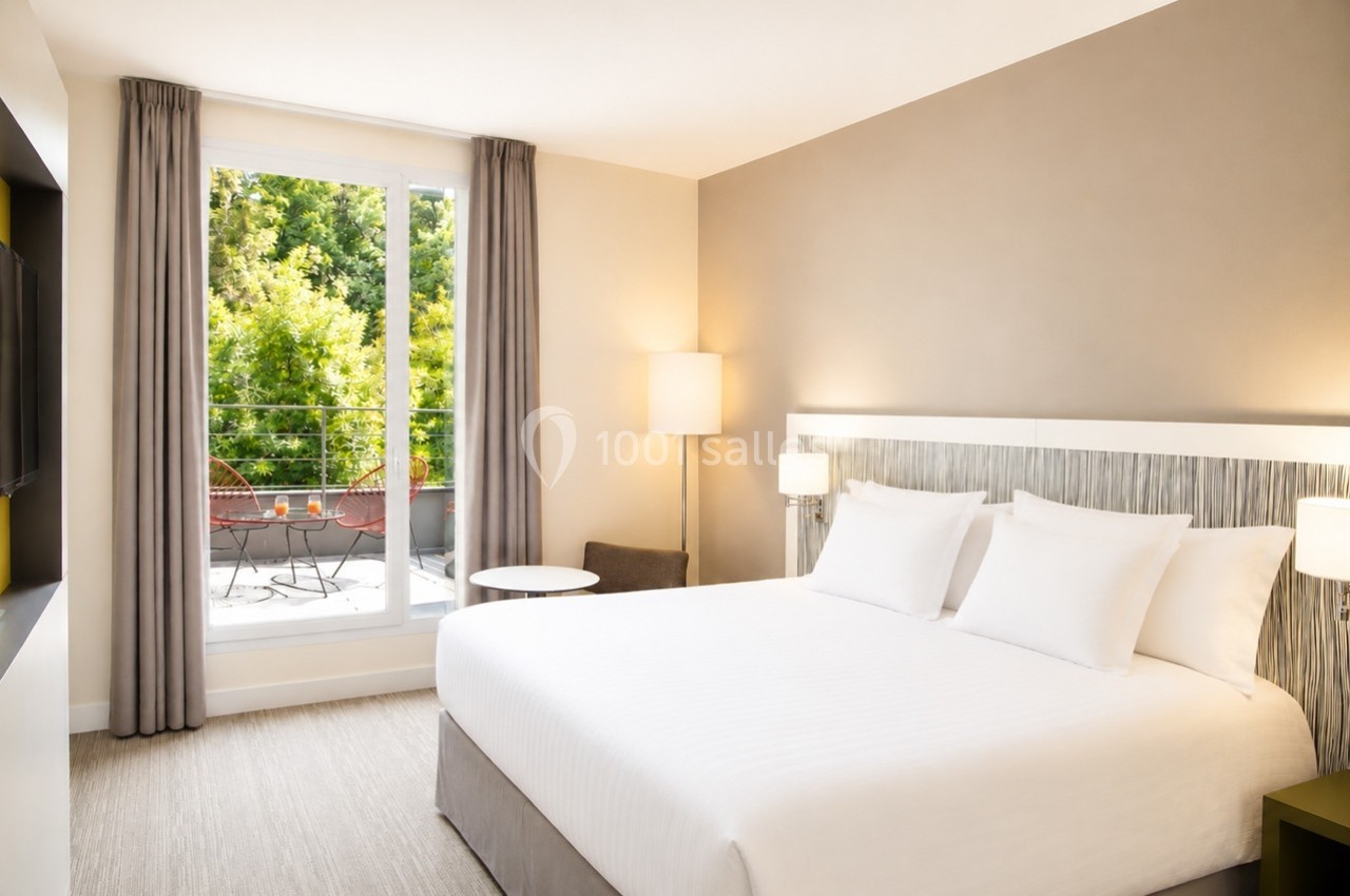 Chambre lumineuse avec un grand lit, des rideaux gris, une table basse et une vue sur un balcon avec végétation.