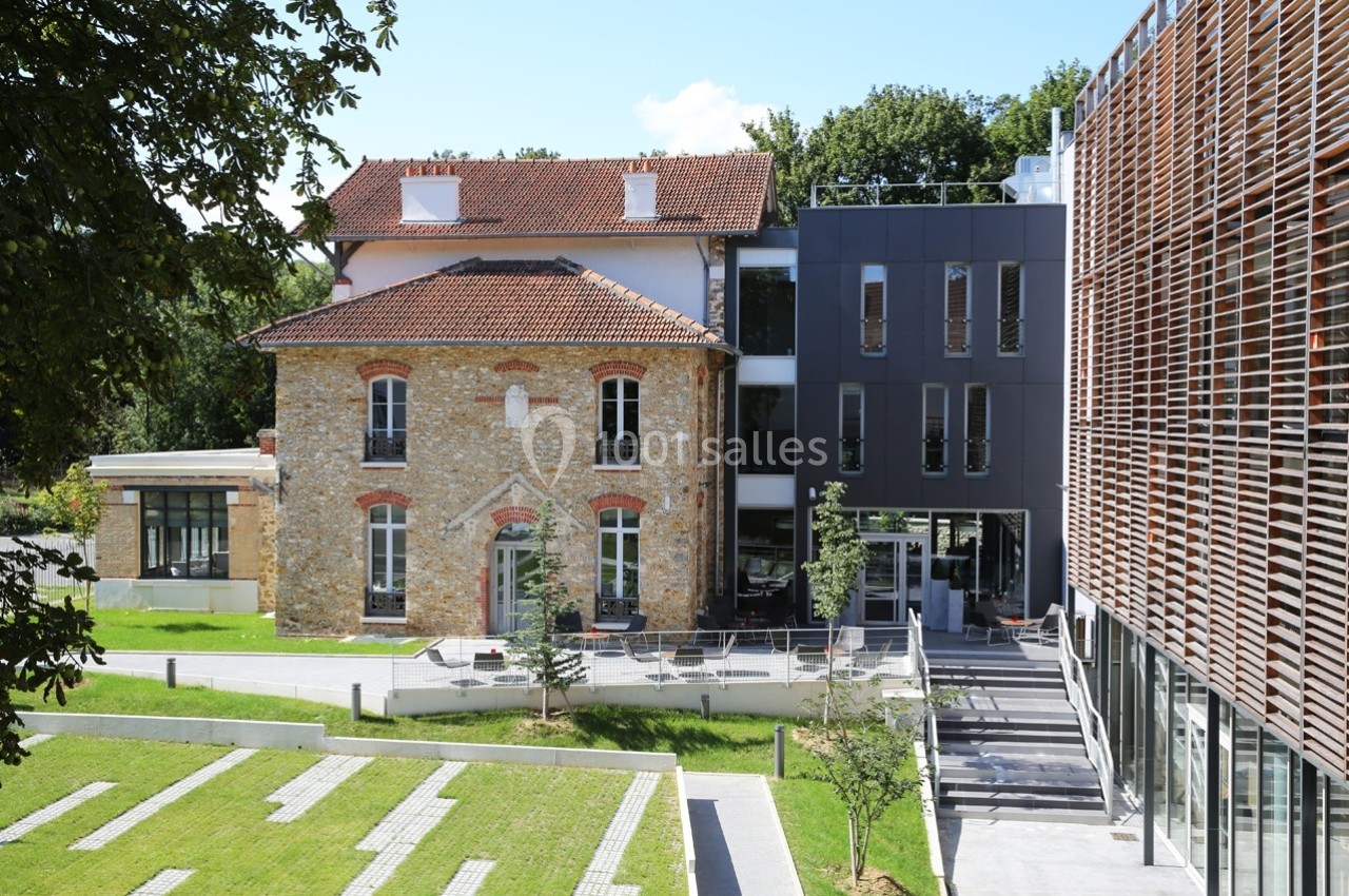 Bâtiment ancien en pierre rénové, adjacent à une structure moderne, entouré d'espaces verts et d'allées pavées.
