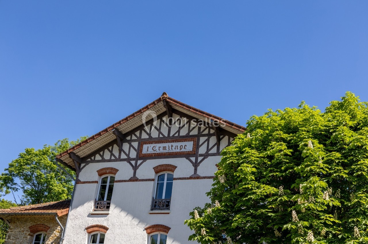 Façade d'une maison à colombages avec l'inscription ’L'Ermitage’, entourée d'arbres sous un ciel bleu.