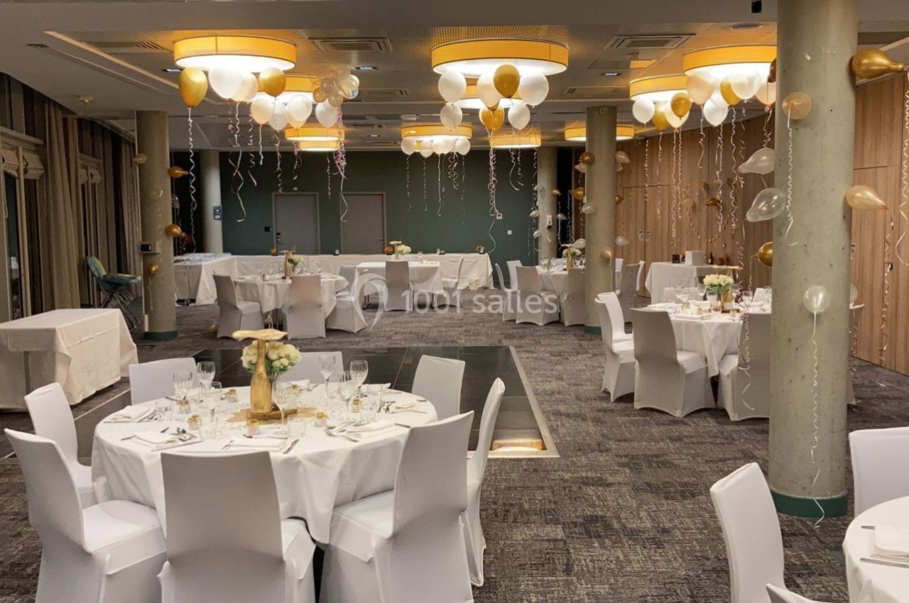 Salle de réception décorée avec des ballons dorés et blancs, tables rondes dressées avec nappes blanches et centres de table.