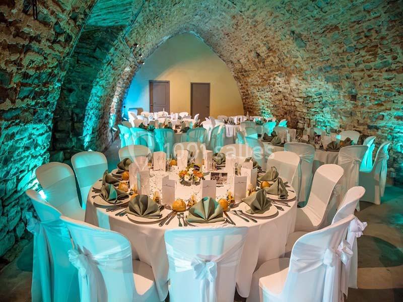 Salle voûtée en pierre décorée pour un événement, avec tables rondes dressées et chaises blanches ornées de nœuds.