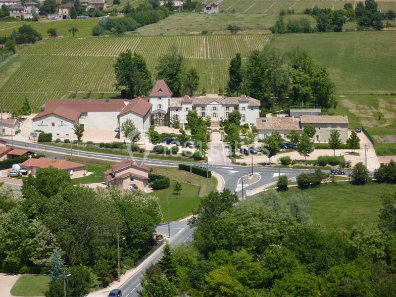 Vue aérienne d'un village avec des bâtiments anciens, des routes, des arbres et des vignobles en arrière-plan.