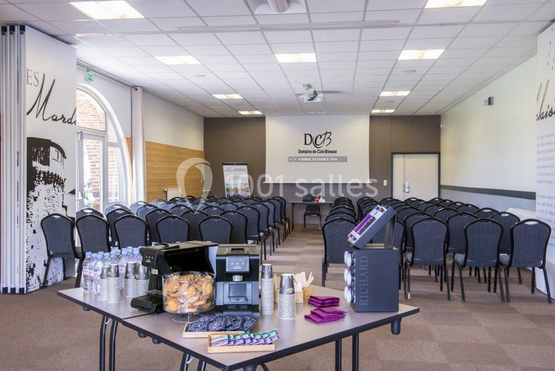 Salle de conférence avec rangées de chaises, table de rafraîchissements au premier plan et écran de présentation au fond.