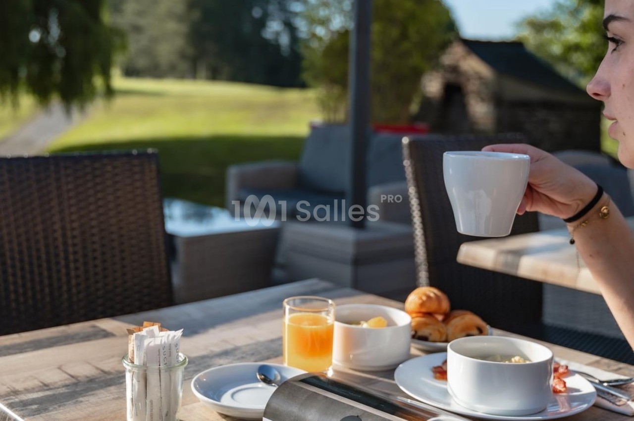 Une personne prend un café en terrasse avec un petit-déjeuner composé de viennoiseries, jus d'orange et confiture.