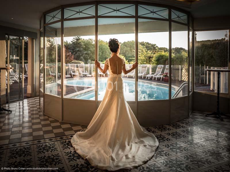 Une femme en robe de mariée regarde une piscine extérieure à travers de grandes baies vitrées.