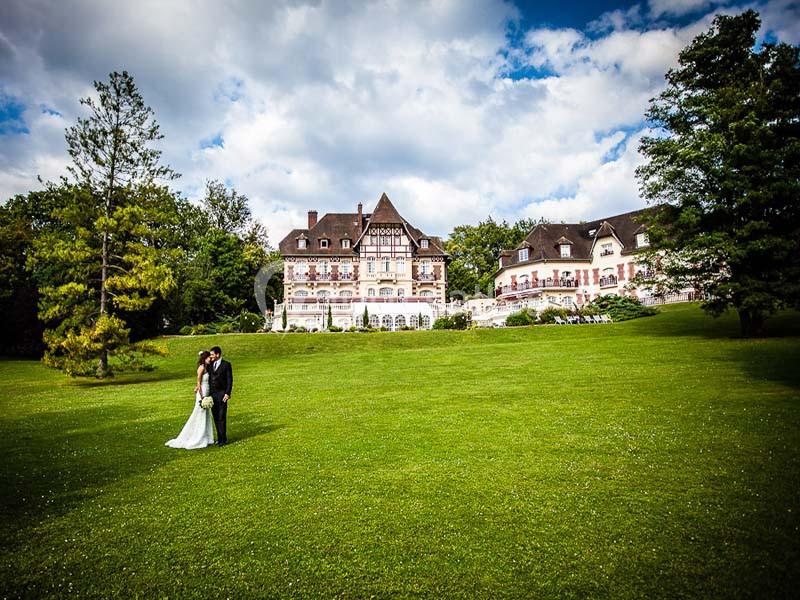 Un couple en tenue de mariage pose sur une pelouse devant un grand bâtiment de style traditionnel entouré d'arbres.
