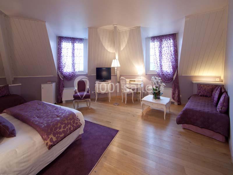 Chambre lumineuse avec parquet, lit double, canapé, bureau, télévision et rideaux violets dans une ambiance élégante.