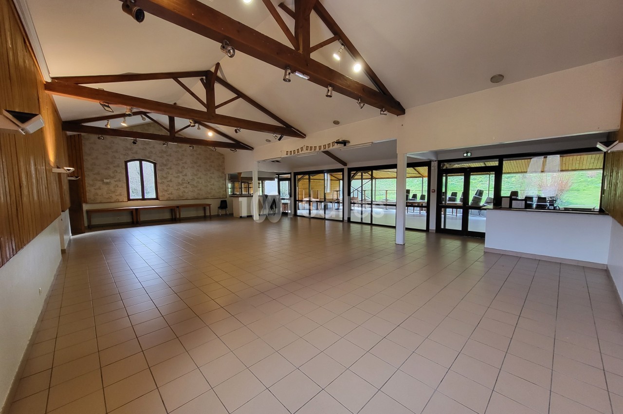 Salle spacieuse avec plafond en poutres apparentes, carrelage au sol et grandes baies vitrées donnant sur l'extérieur.