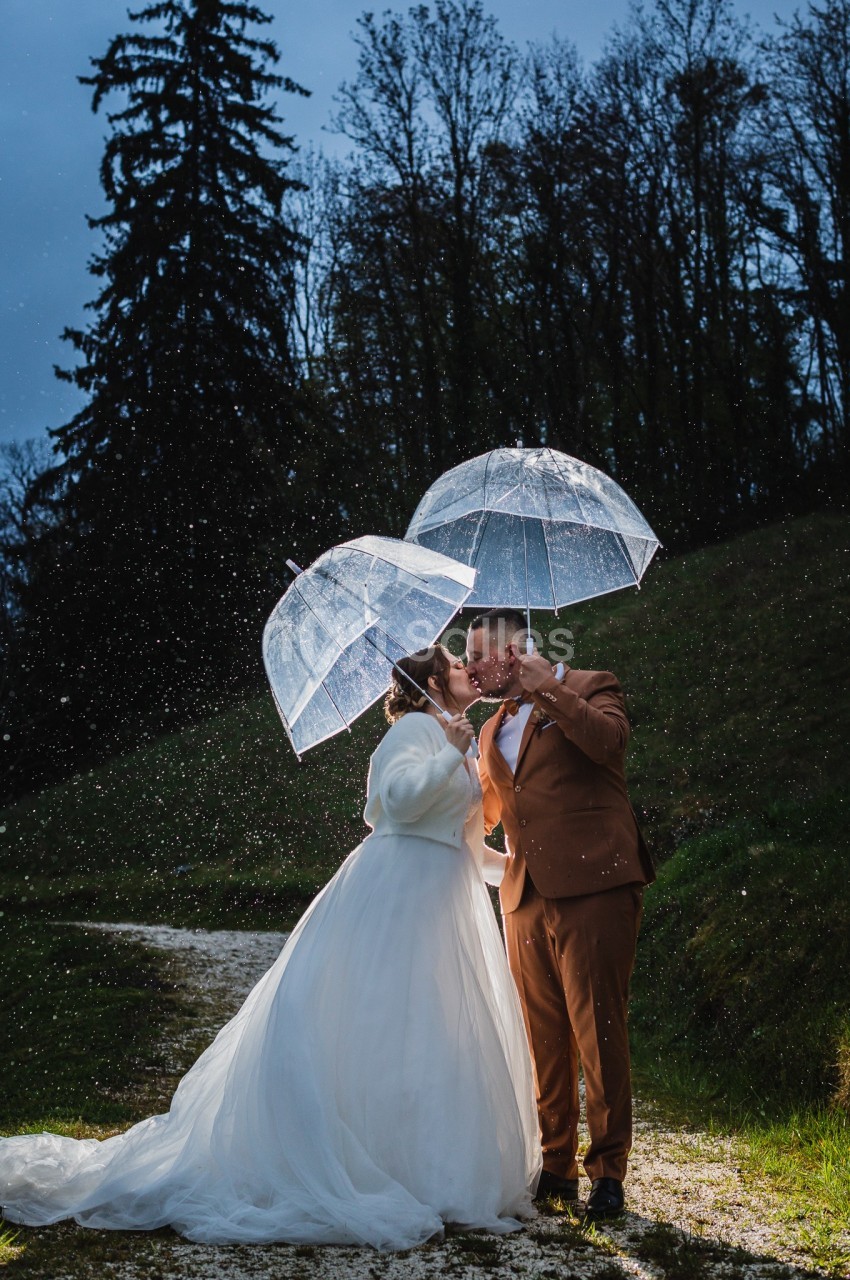 Un couple sous la pluie partage un baiser, abrité par des parapluies transparents dans un paysage naturel en soirée.