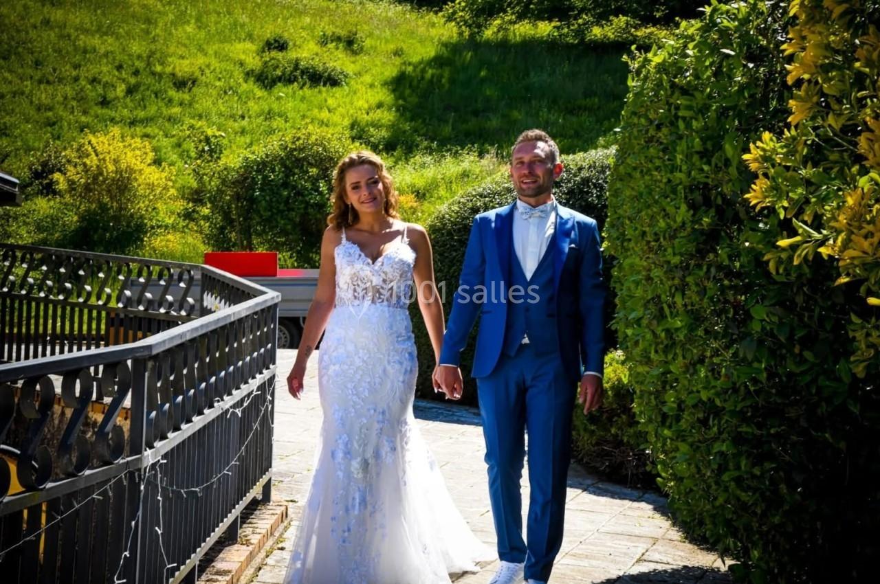 Un couple en tenue de mariage marche sur un chemin bordé de verdure par une journée ensoleillée.