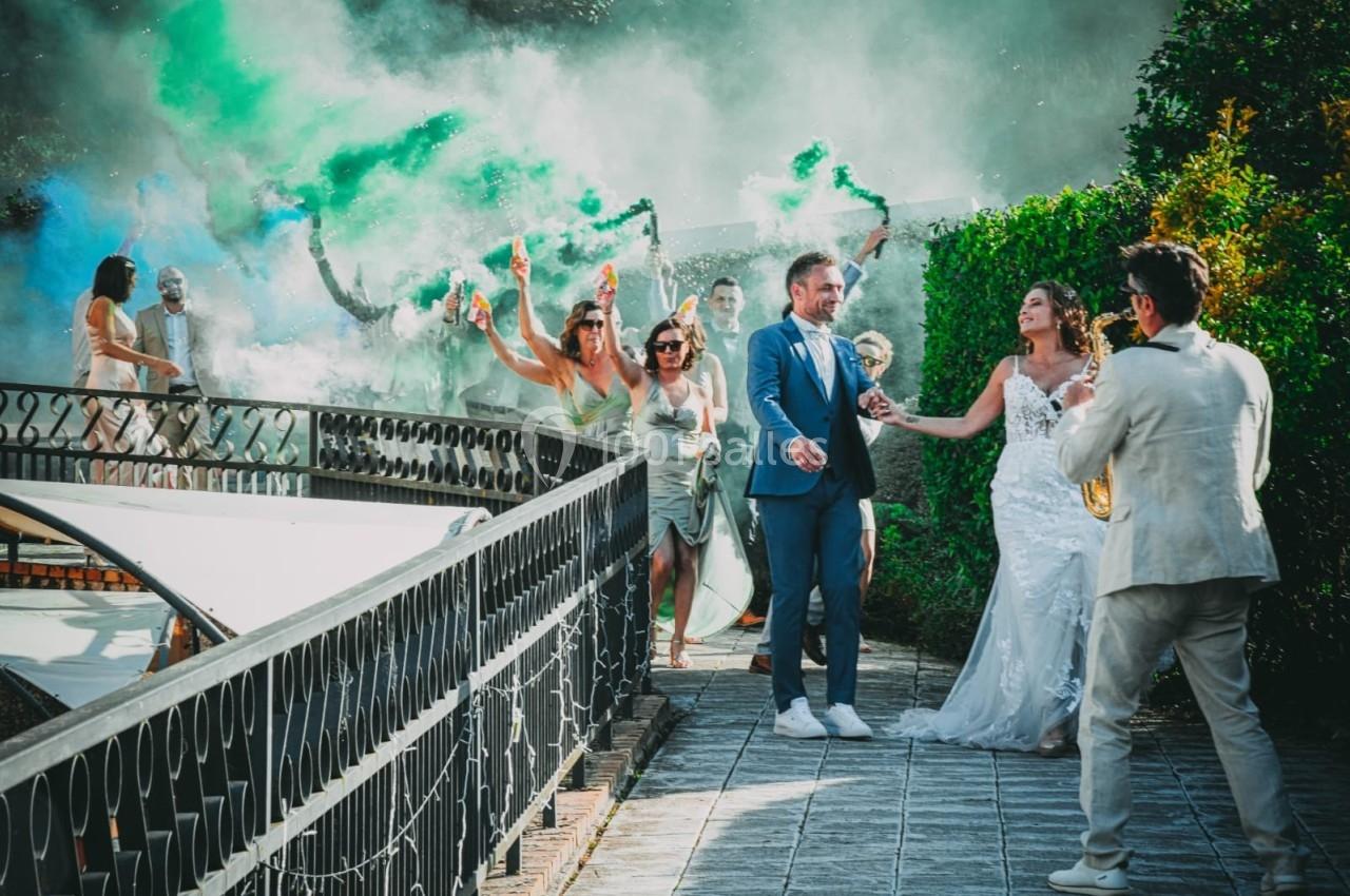 Un couple en tenue de mariage marche sur une allée tandis que des invités célèbrent avec des fumigènes colorés.