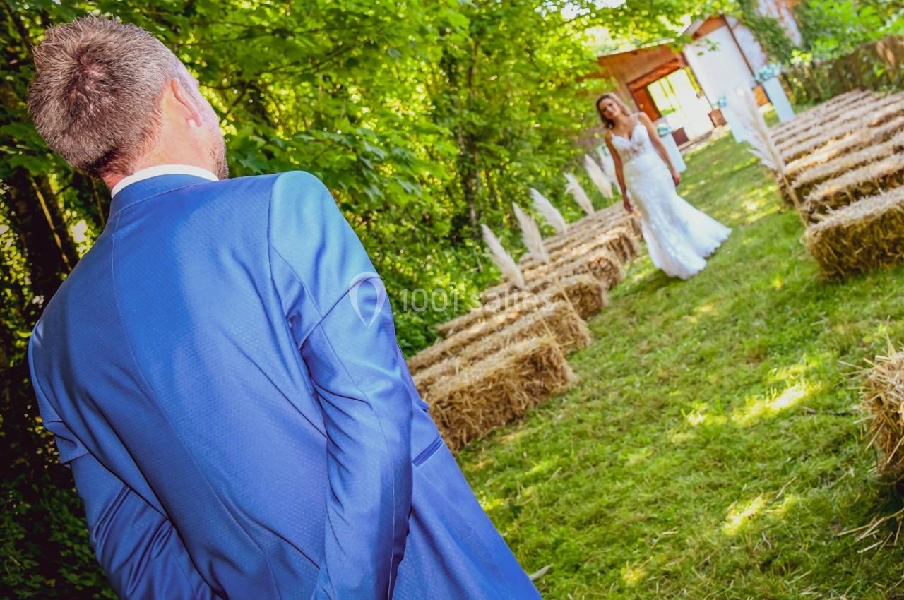 Un homme en costume bleu regarde une femme en robe de mariée avançant dans une allée bordée de bottes de paille.