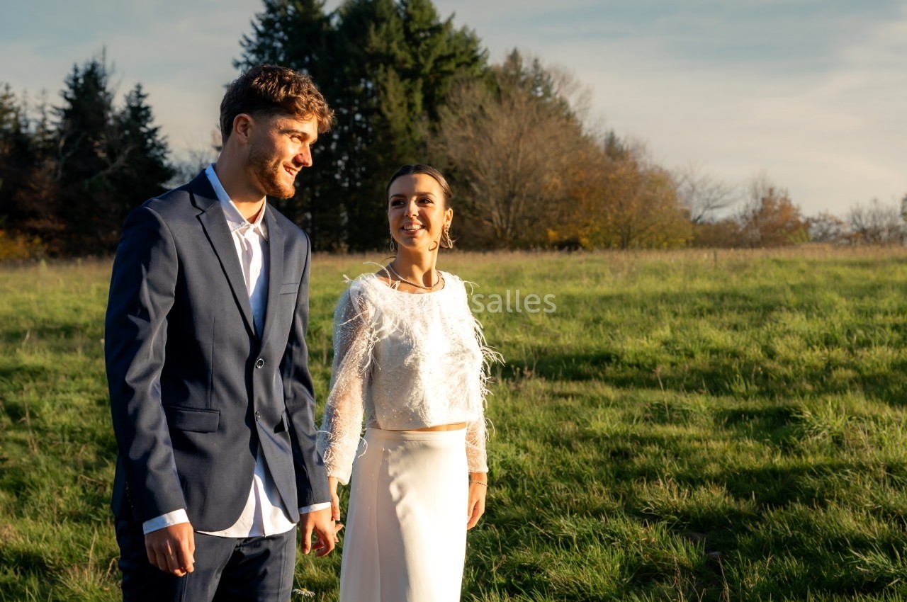 Un couple en tenue élégante marche dans un champ verdoyant sous une lumière douce de fin de journée.