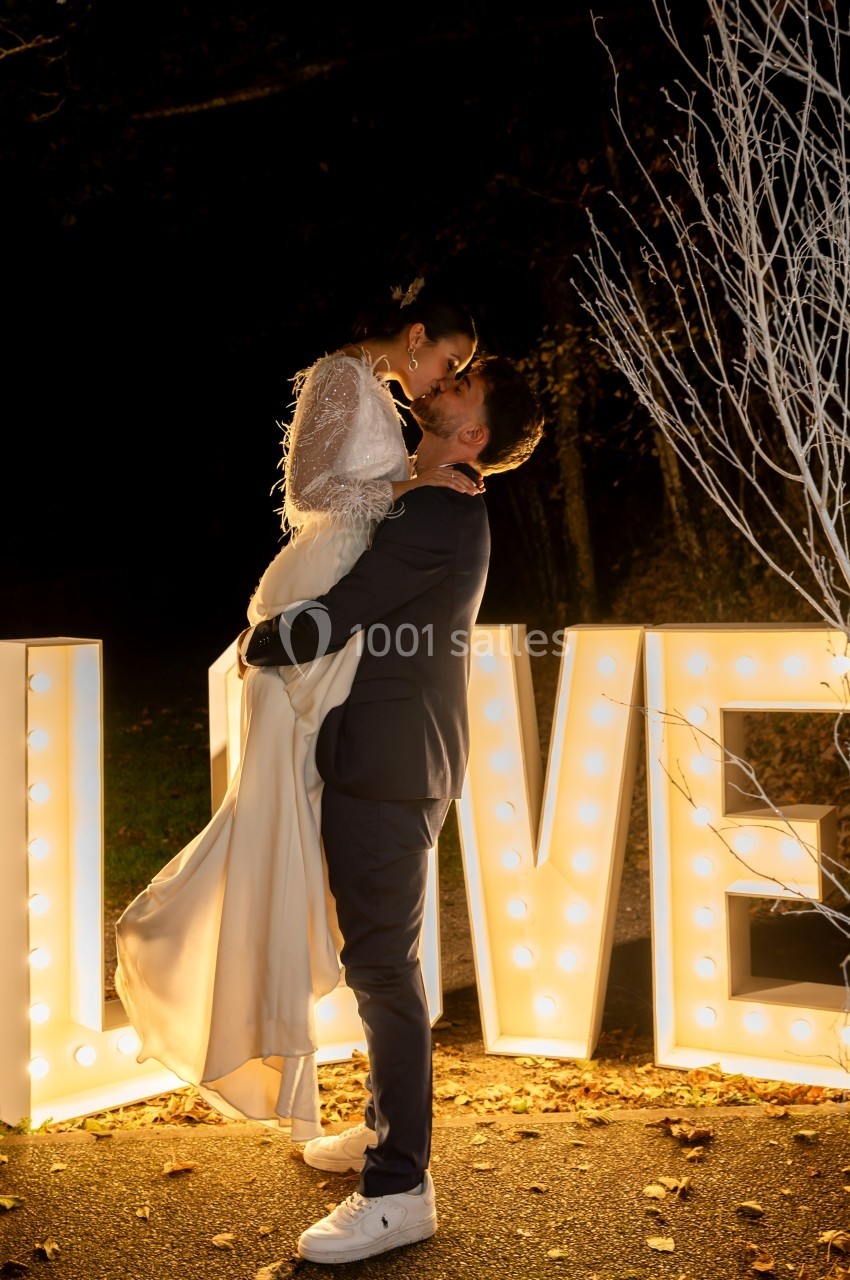 Un couple en tenue de mariage s'enlace devant des lettres lumineuses formant le mot ’LOVE’ dans un décor nocturne.