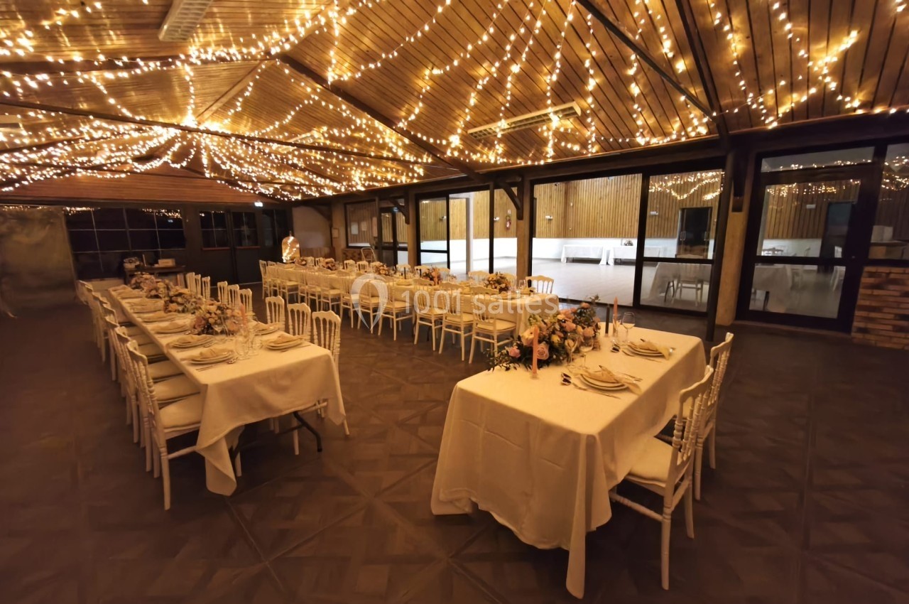 Salle décorée pour un événement avec tables dressées, nappes blanches et guirlandes lumineuses suspendues au plafond.