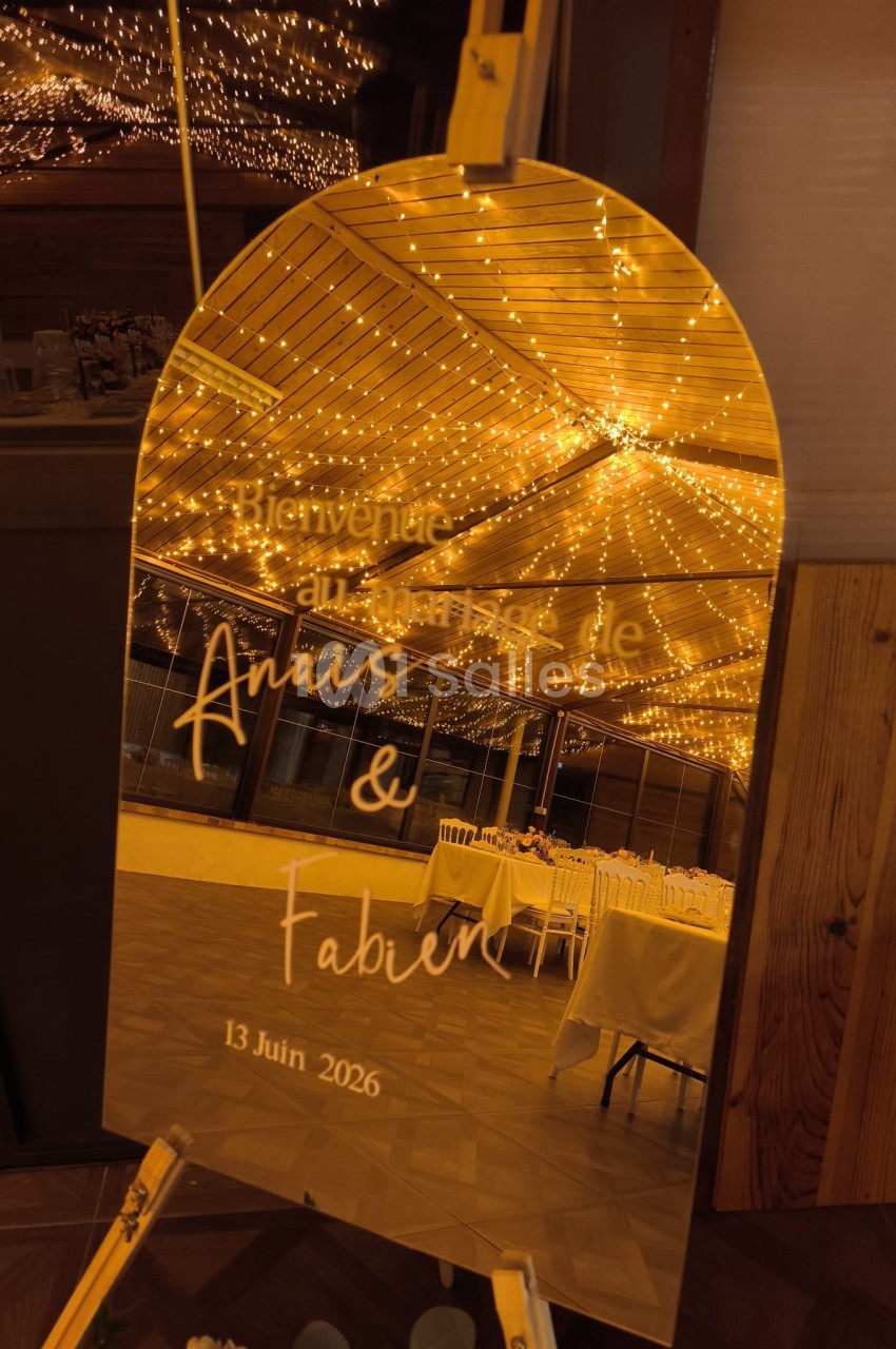 Miroir doré avec inscription personnalisée pour un mariage, reflétant une salle décorée de guirlandes lumineuses.