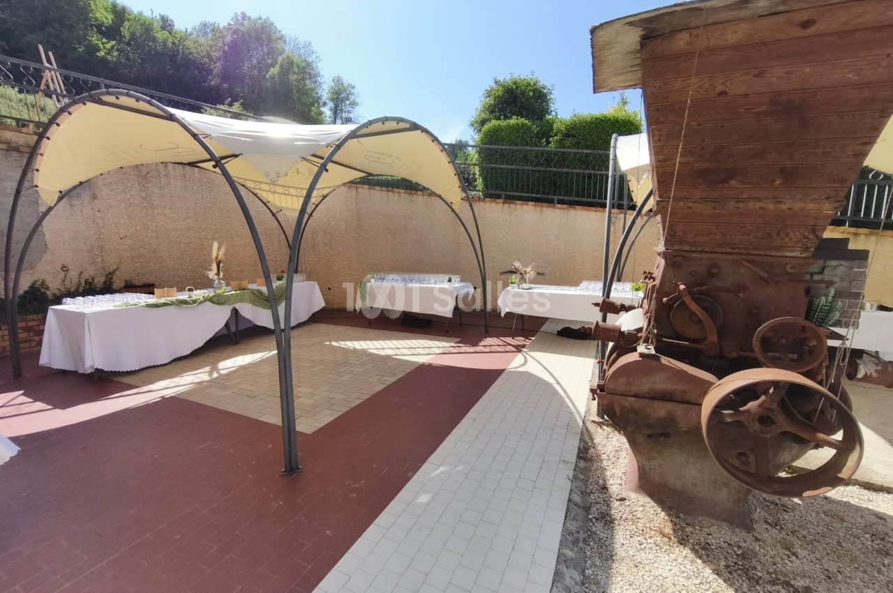 Espace extérieur avec tables dressées sous des tonnelles, près d'une ancienne machine agricole en bois et métal.