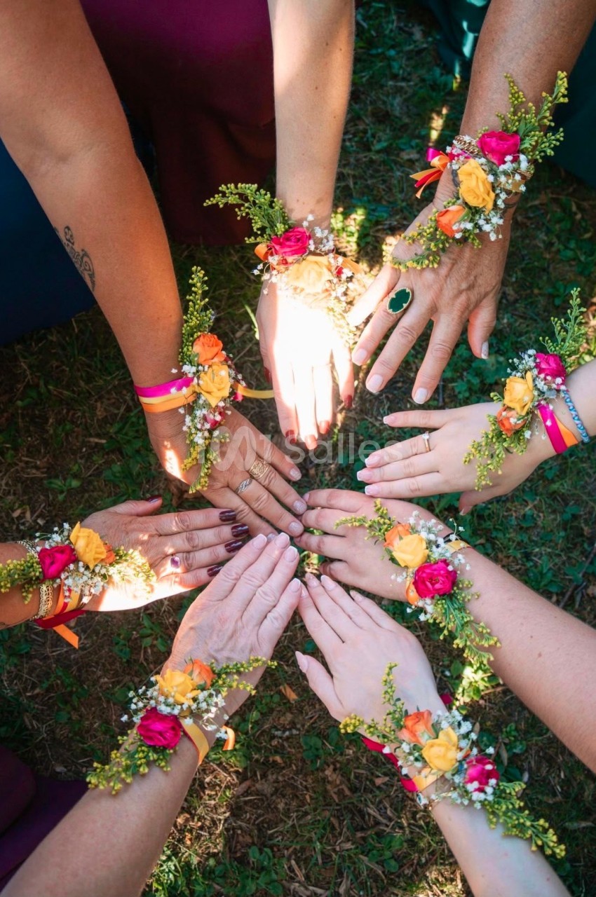 Mains de plusieurs personnes portant des bracelets fleuris, disposées en cercle sur de l'herbe.