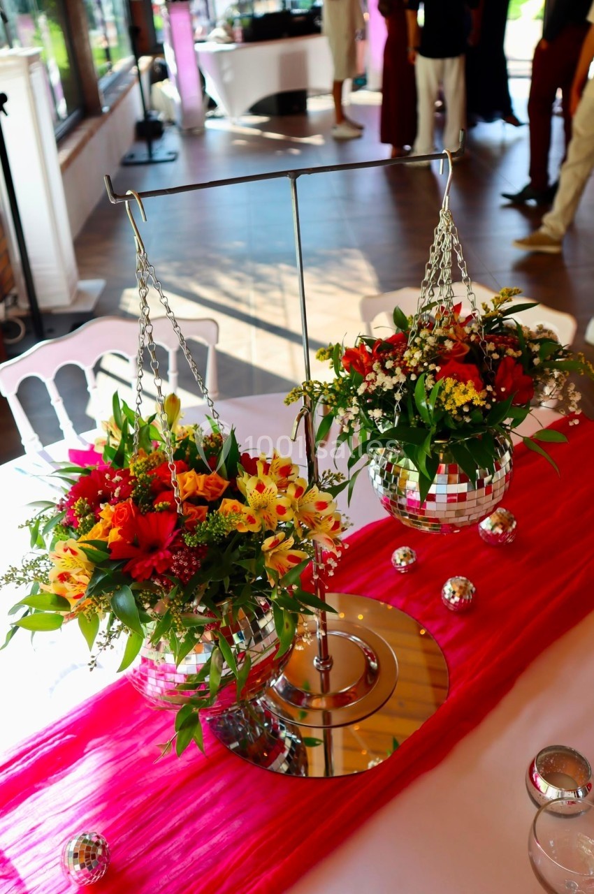 Deux compositions florales colorées suspendues à des boules miroir sur une table décorée d'un tissu rose.