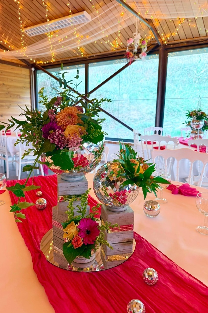 Centre de table coloré avec fleurs, boules à facettes et nappe rouge, dans une salle décorée pour un événement festif.