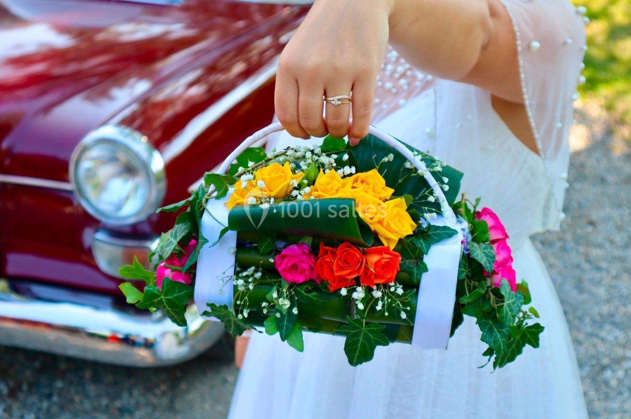 Une main tenant un panier de fleurs colorées devant une voiture ancienne rouge.