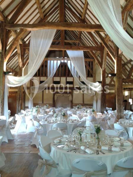 Salle de réception rustique avec poutres apparentes, tables rondes dressées et voilages suspendus.