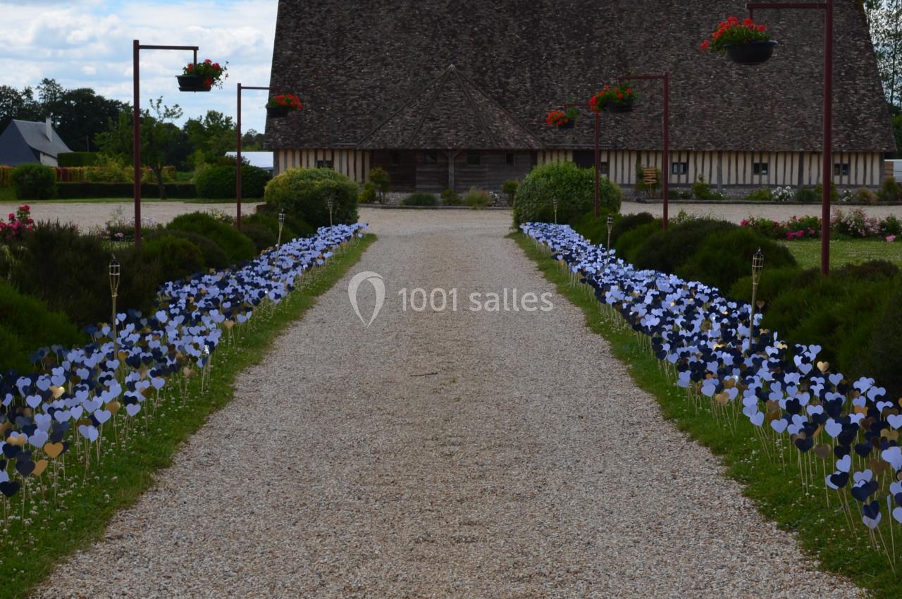 Allée gravillonnée bordée de fleurs décoratives menant à une maison à colombages sous un ciel partiellement nuageux.