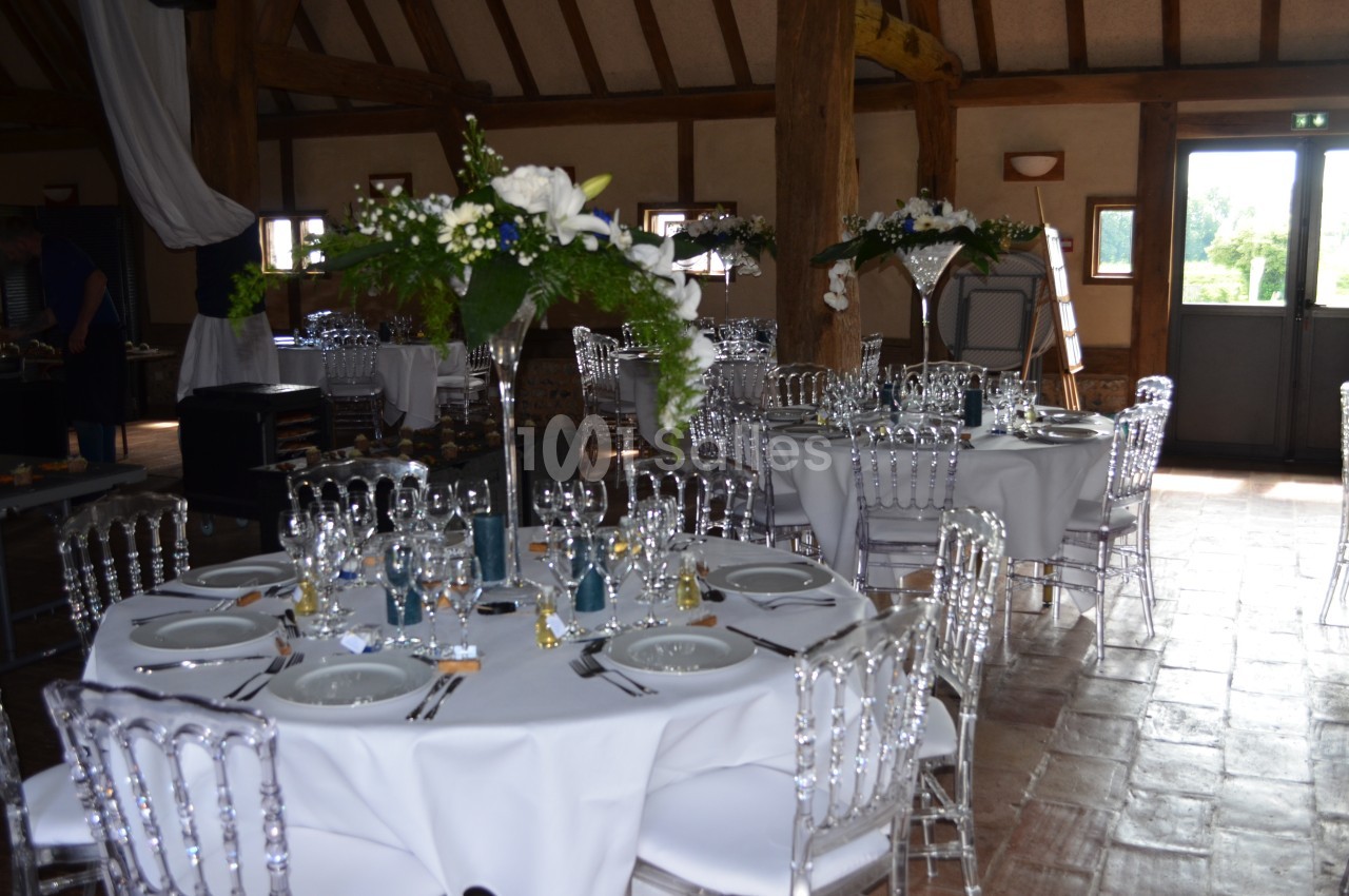 Salle de réception avec tables rondes dressées, chaises transparentes et décorations florales blanches et vertes.