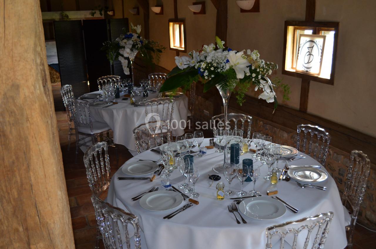 Salle rustique décorée pour un événement, avec tables rondes dressées, chaises transparentes et compositions florales.