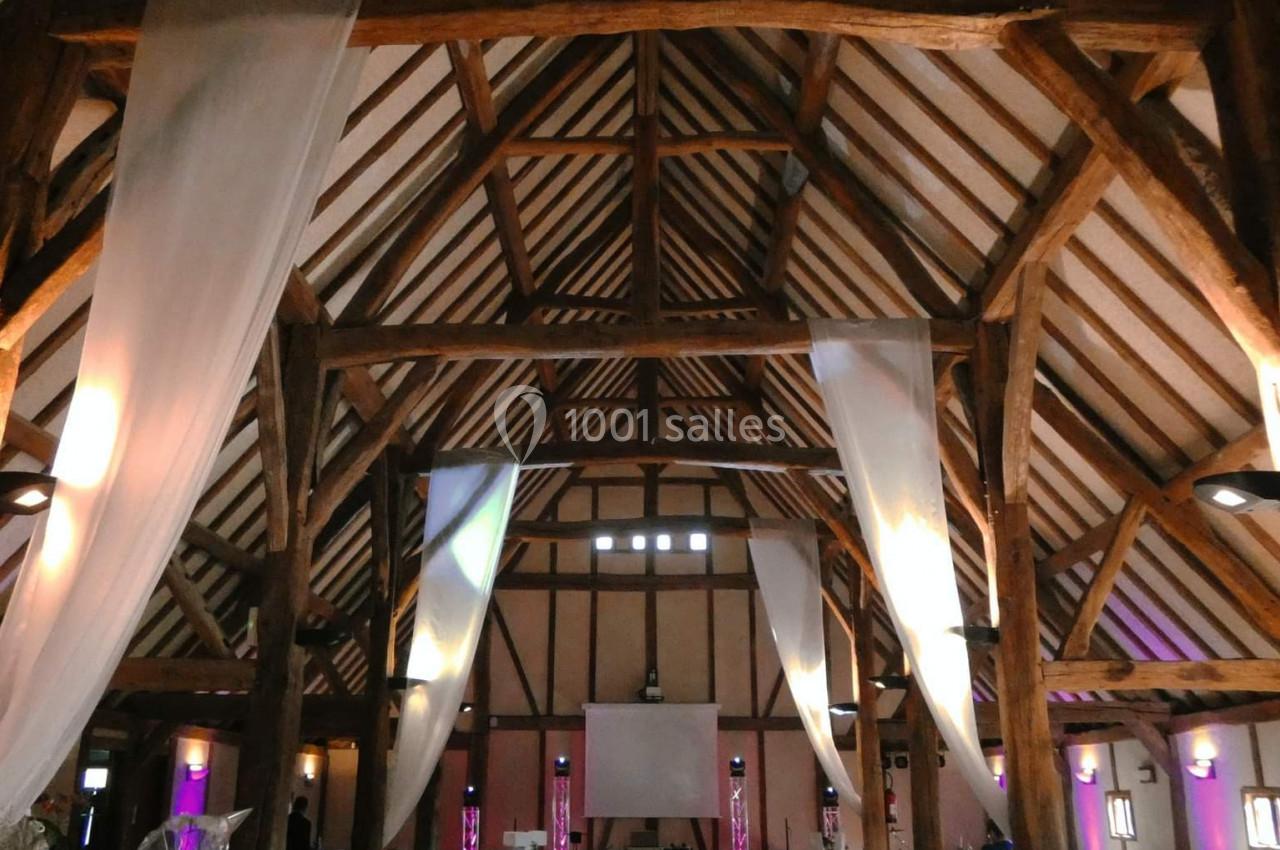 Grande salle de réception avec charpente en bois apparente, décorée de voilages blancs et éclairages colorés.