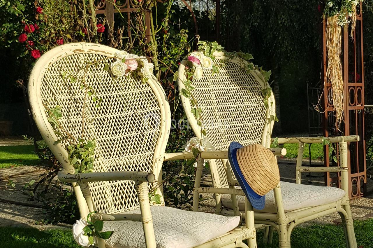 Deux chaises en osier décorées de fleurs, placées sur une pelouse devant une arche métallique ornée de végétation.