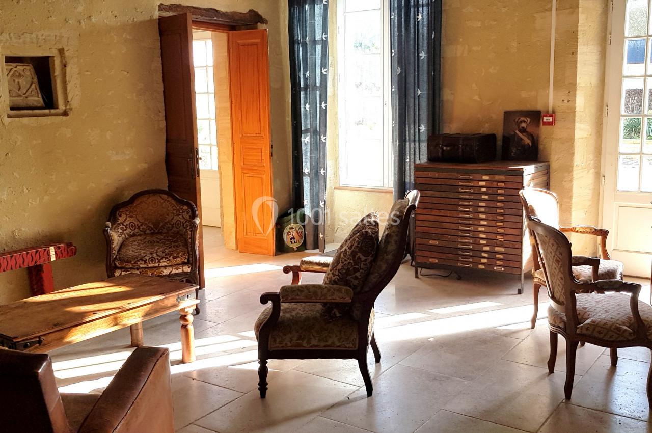 Salon lumineux avec mobilier ancien, fauteuils rembourrés, table en bois et fenêtres donnant sur un jardin.
