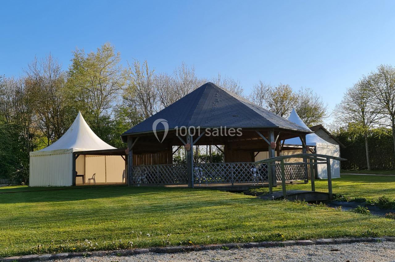 Abri en bois avec toit en ardoise entouré de tentes blanches, situé dans un espace vert avec arbres en arrière-plan.